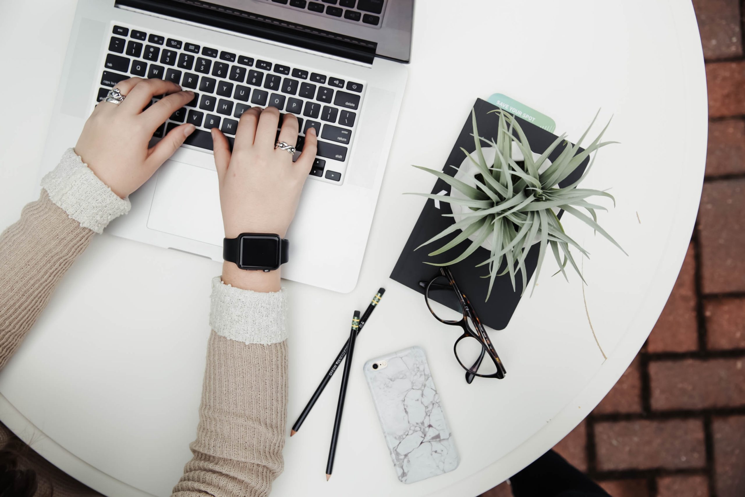 a ladies hands on a laptop keyboard, next to her are black glasses, a marble phone case and a succulent plant. The view is from above